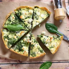 Pyszna tarta ze szpinakiem i fetą, pokrojona w trójkąty, leży na papierze do pieczenia. Obok nóż i świeże liście szpinaku.