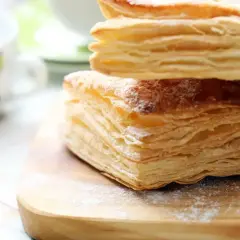 Stos warstw ciasta francuskiego, idealny do nauki jak zrobić ciasto francuskie. Posypane cukrem pudrem, obok filiżanki.