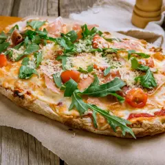 Pyszna pinsa z szynką parmeńską, rukolą i pomidorkami koktajlowymi, posypana parmezanem.