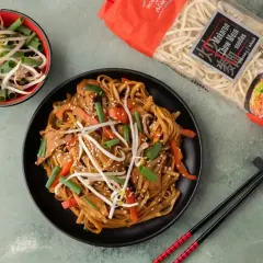 Gotowy do jedzenia makaron chow mein z warzywami i kiełkami fasoli. Obok opakowanie makaronu.
