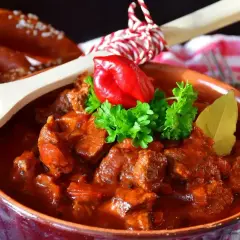 Aromatyczny gulasz z kurczaka w glinianej misie, ozdobiony papryczką chili i natką pietruszki. Obok pieczywo i drewniana łyżka z pieprzem.