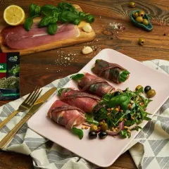 Roladki z szynką parmeńską i anchois, podane z rukolą, oliwkami i orzeszkami pinii. Obok talerza złote sztućce i opakowanie tymianku.