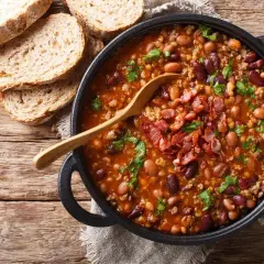 Gorąca fasolka po bretońsku w garnku, posypana boczkiem i natką pietruszki, obok kromki chleba.