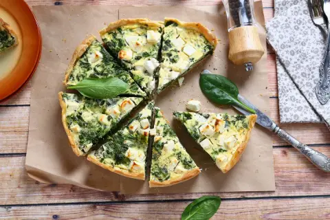 Pyszna tarta ze szpinakiem i fetą, pokrojona w trójkąty, leży na papierze do pieczenia. Obok nóż i świeże liście szpinaku.