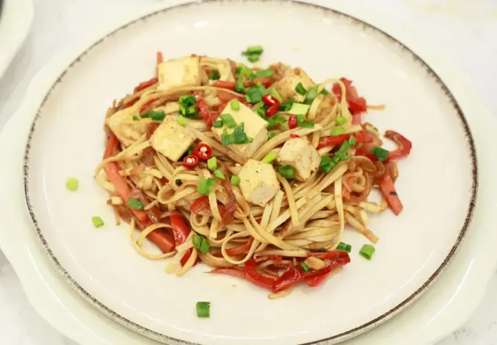 Pyszny makaron z tofu, papryką i szczypiorkiem, posypany chili. Danie wygląda apetycznie i kolorowo.