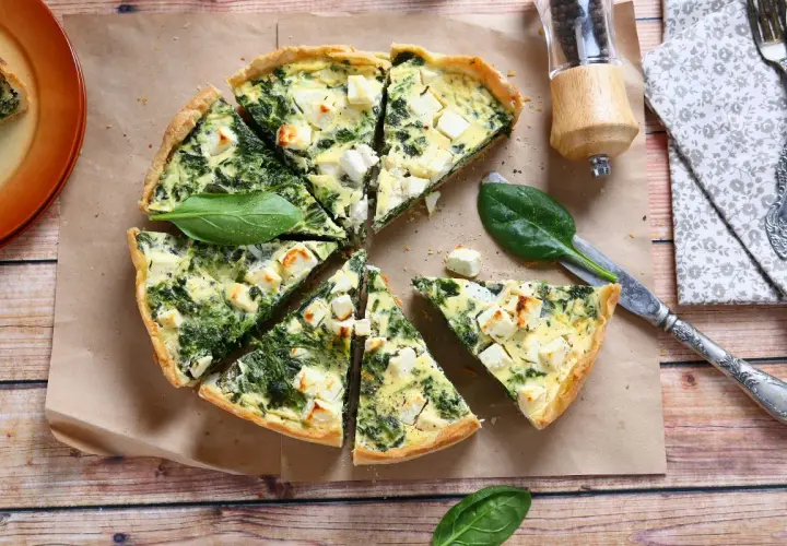 Pyszna tarta ze szpinakiem i fetą, pokrojona w trójkąty, leży na papierze do pieczenia. Obok nóż i świeże liście szpinaku.