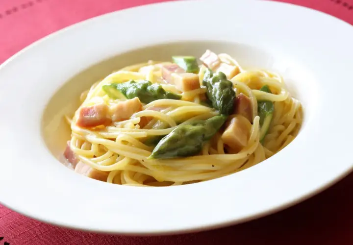 Pyszna carbonara ze szparagami, podana w białej misce na czerwonym tle.