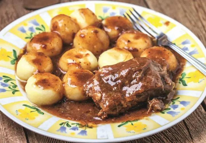 Mięciutka karkówka w sosie, podana z kluskami śląskimi na talerzu z żółto-białym wzorem.