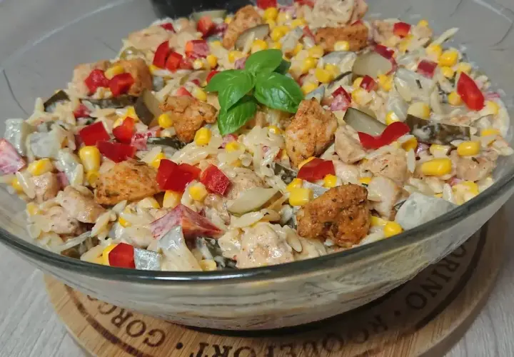 Pyszna sałatka z kurczakiem, makaronem orzo, kukurydzą, papryką i ogórkiem kiszonym, udekorowana listkiem bazylii.