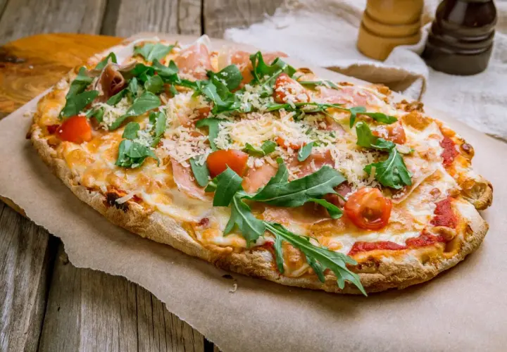Pyszna pinsa z szynką parmeńską, rukolą i pomidorkami koktajlowymi, posypana parmezanem.