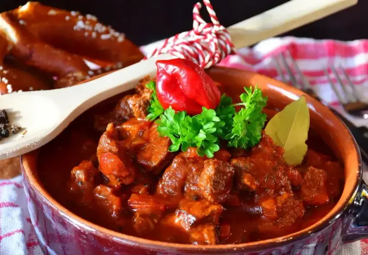 Aromatyczny gulasz z kurczaka w glinianej misie, ozdobiony papryczką chili i natką pietruszki. Obok pieczywo i drewniana łyżka z pieprzem.