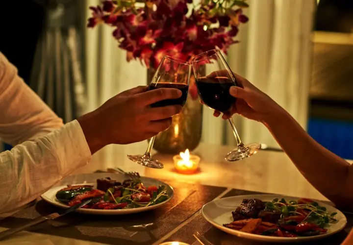 Romantyczna kolacja dla dwojga przy świecach. Dwie pary kieliszków z czerwonym winem unoszą się w powietrzu, a na talerzach czekają pyszne dania.