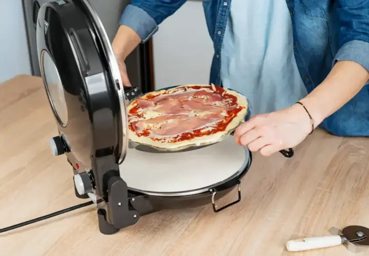 Osoba wkłada pizzę z szynką do pieca. Szybko i łatwo przygotujesz idealną pizzę w domu.