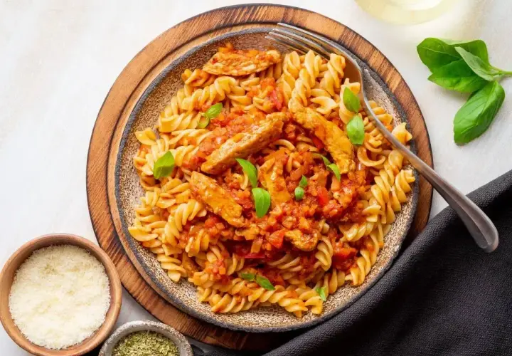 Pyszny makaron z kurczakiem w sosie pomidorowym, udekorowany bazylią. Obok miseczki z parmezanem i ziołami.