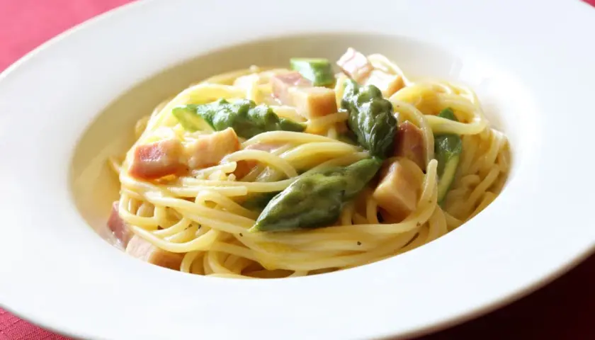 Pyszna carbonara ze szparagami, podana w białej misce na czerwonym tle.