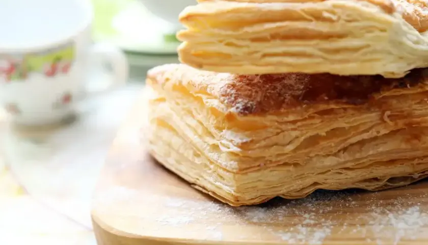 Stos warstw ciasta francuskiego, idealny do nauki jak zrobić ciasto francuskie. Posypane cukrem pudrem, obok filiżanki.
