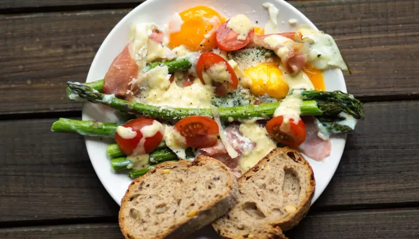 Pyszne szparagi z szynką parmeńską, jajkiem sadzonym i pomidorkami, polane sosem. Podane z chlebem.