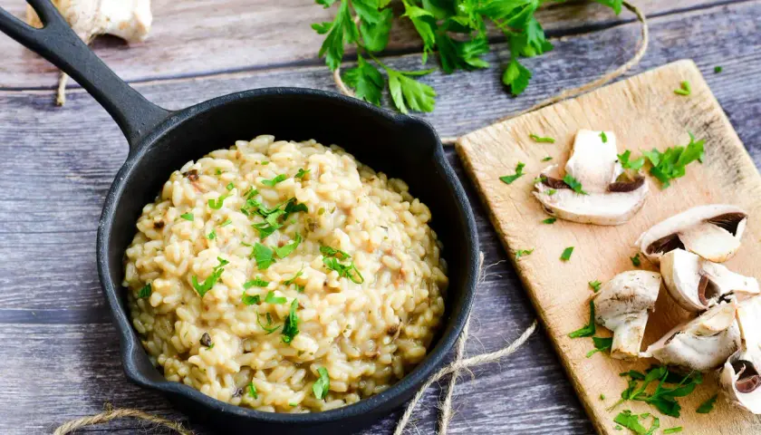 Kremowe risotto z warzywami, posypane natką pietruszki, obok świeże pieczarki na desce.