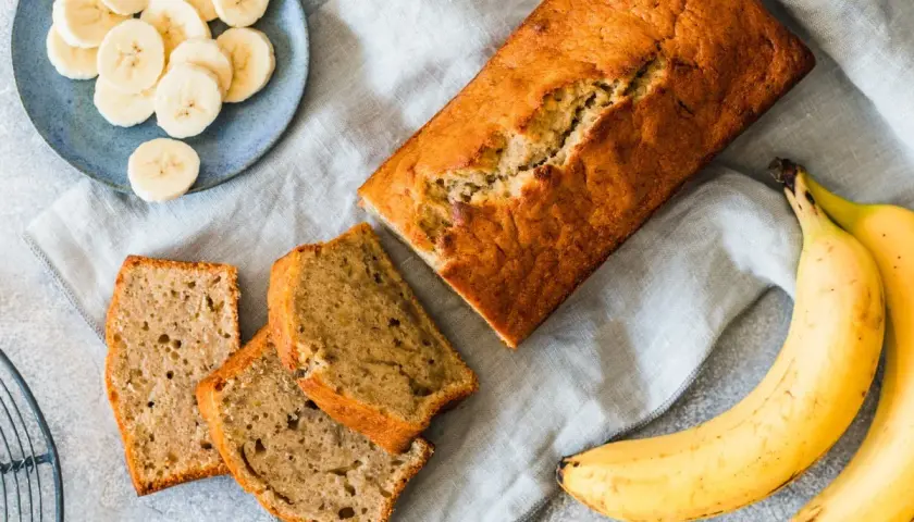 Chlebek bananowy pokrojony na kromki, obok banany i talerzyk z plasterkami banana.