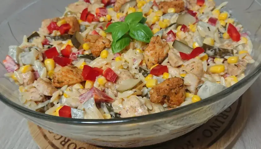 Pyszna sałatka z kurczakiem, makaronem orzo, kukurydzą, papryką i ogórkiem kiszonym, udekorowana listkiem bazylii.