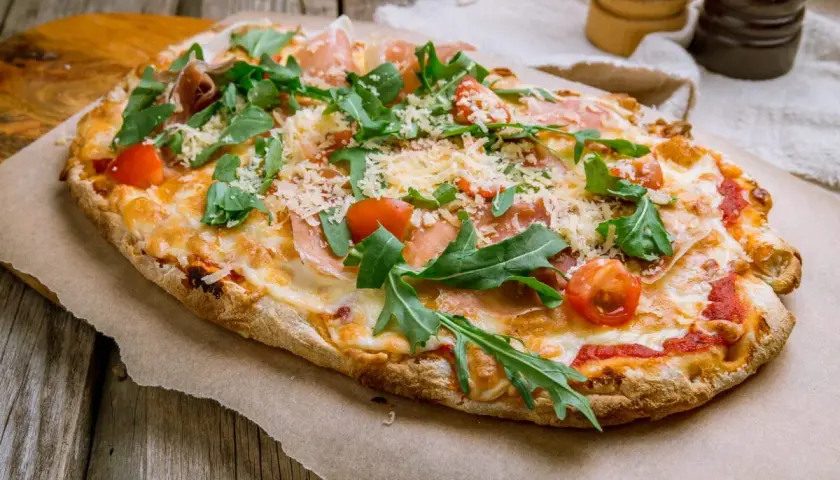 Pyszna pinsa z szynką parmeńską, rukolą i pomidorkami koktajlowymi, posypana parmezanem.