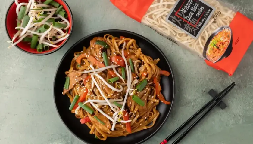 Gotowy do jedzenia makaron chow mein z warzywami i kiełkami fasoli. Obok opakowanie makaronu.