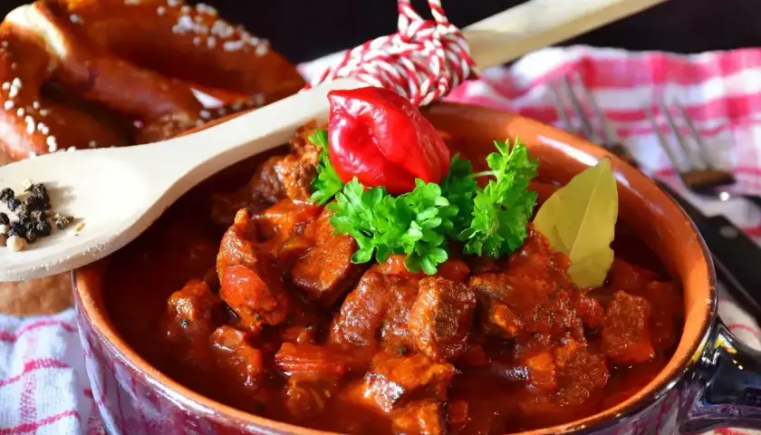 Aromatyczny gulasz z kurczaka w glinianej misie, ozdobiony papryczką chili i natką pietruszki. Obok pieczywo i drewniana łyżka z pieprzem.