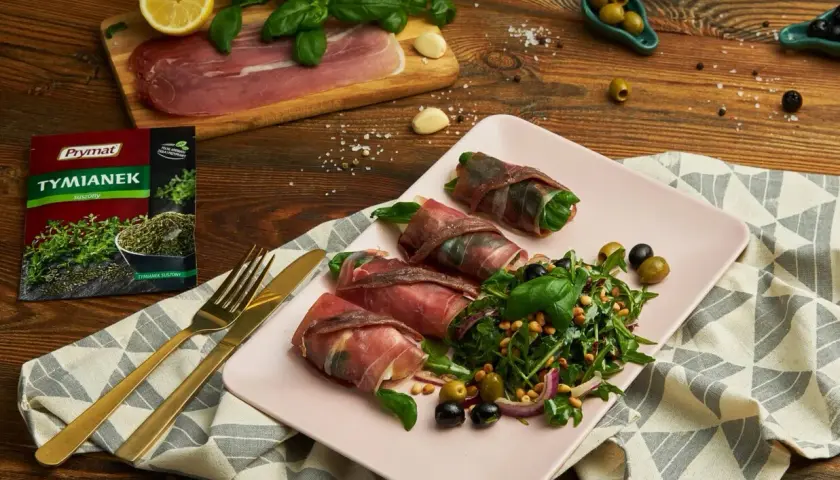Roladki z szynką parmeńską i anchois, podane z rukolą, oliwkami i orzeszkami pinii. Obok talerza złote sztućce i opakowanie tymianku.