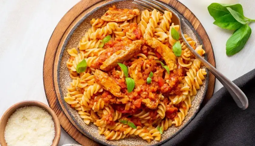 Pyszny makaron z kurczakiem w sosie pomidorowym, udekorowany bazylią. Obok miseczki z parmezanem i ziołami.