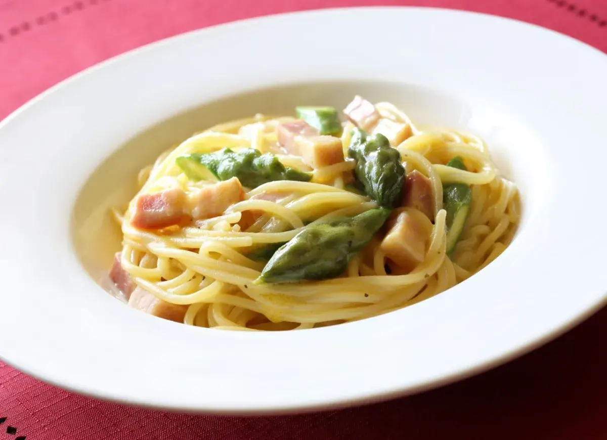 Pyszna carbonara ze szparagami, podana w białej misce na czerwonym tle.