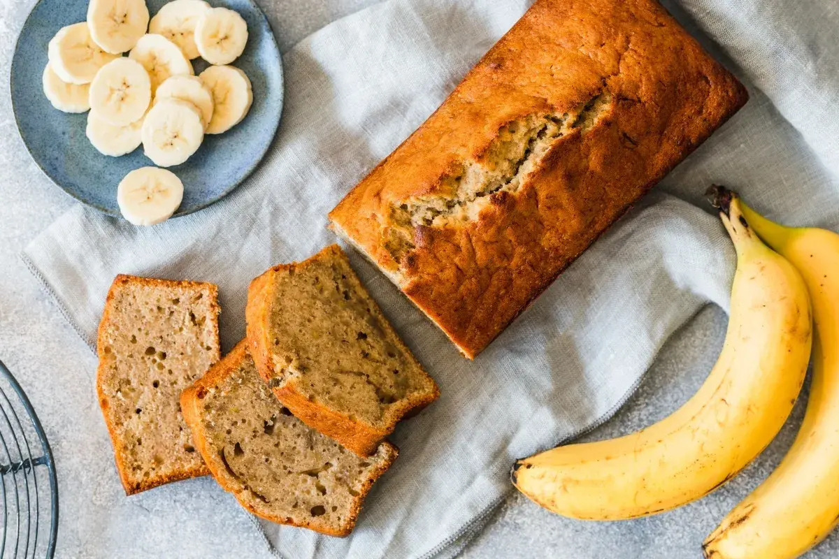 Chlebek bananowy pokrojony na kromki, obok banany i talerzyk z plasterkami banana.
