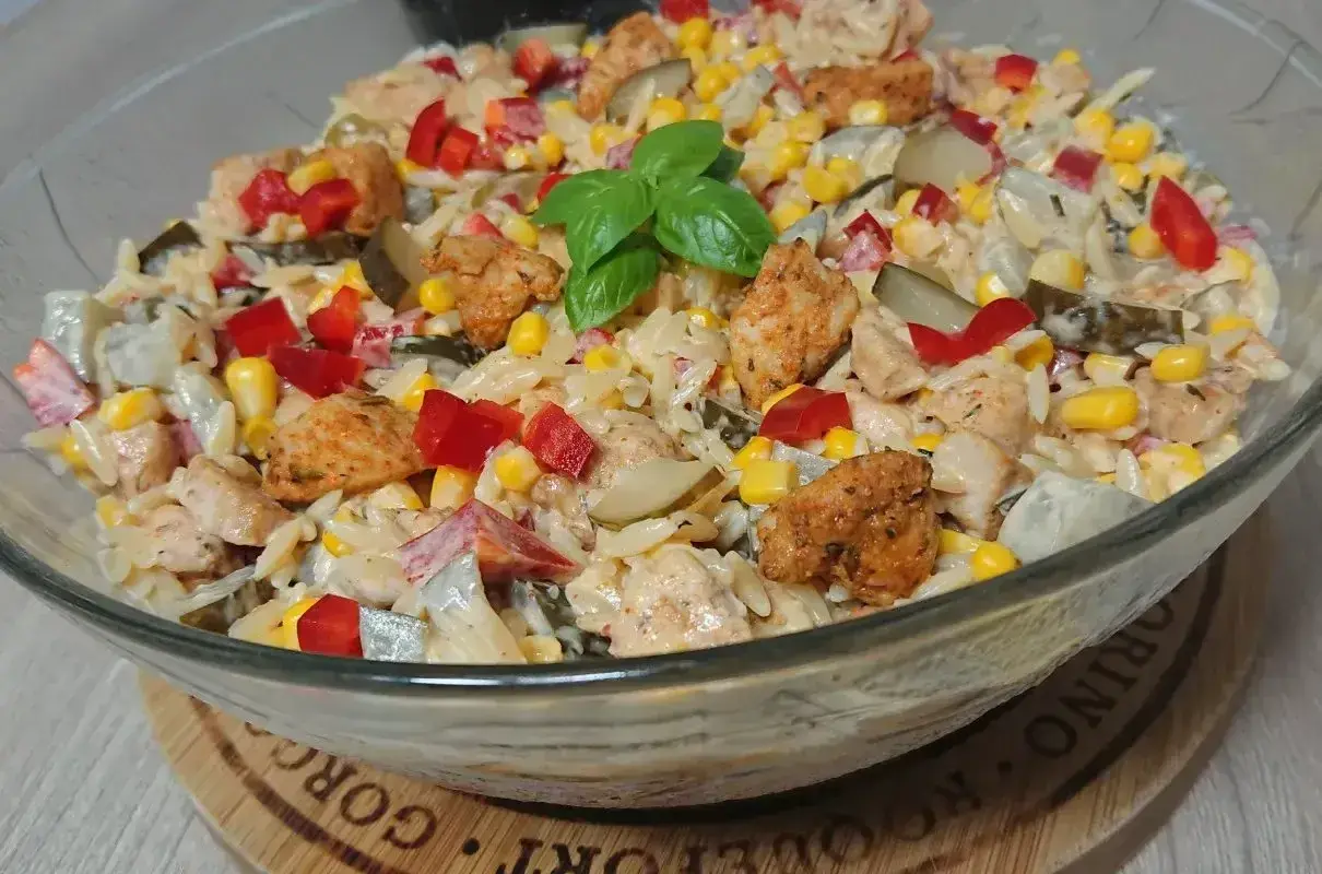 Pyszna sałatka z kurczakiem, makaronem orzo, kukurydzą, papryką i ogórkiem kiszonym, udekorowana listkiem bazylii.
