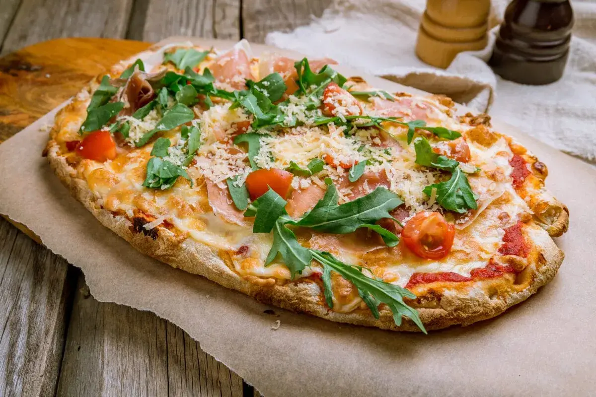 Pyszna pinsa z szynką parmeńską, rukolą i pomidorkami koktajlowymi, posypana parmezanem.