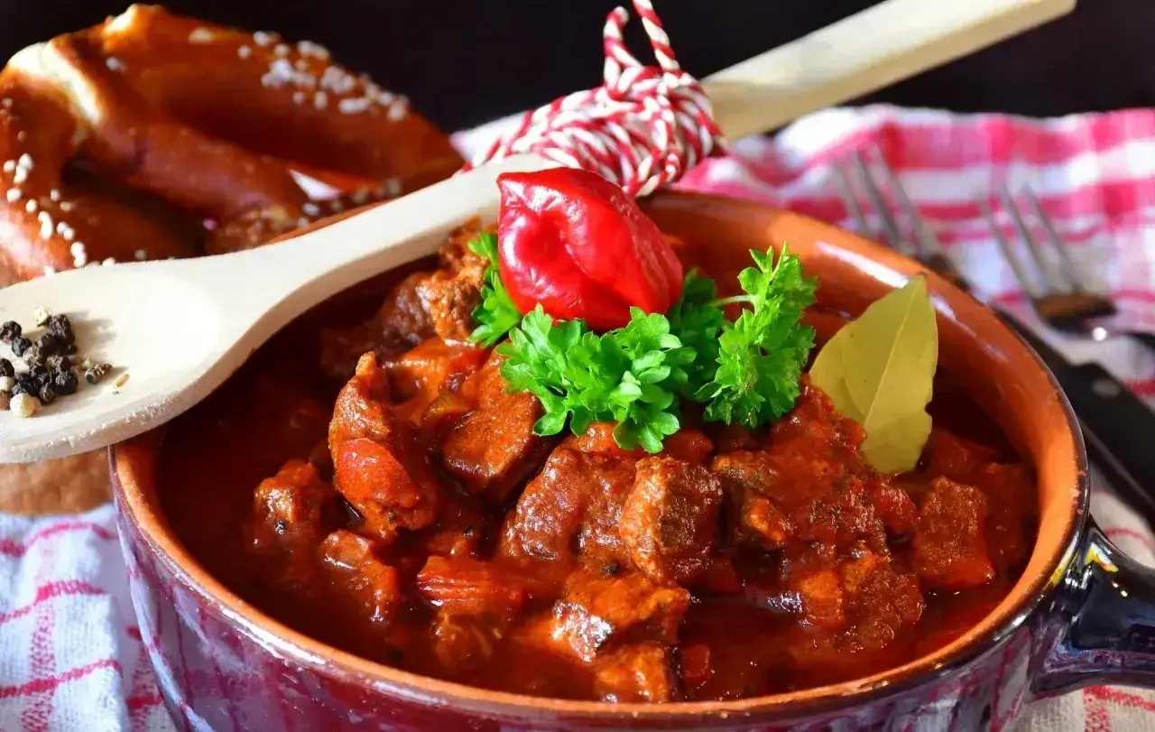 Aromatyczny gulasz z kurczaka w glinianej misie, ozdobiony papryczką chili i natką pietruszki. Obok pieczywo i drewniana łyżka z pieprzem.