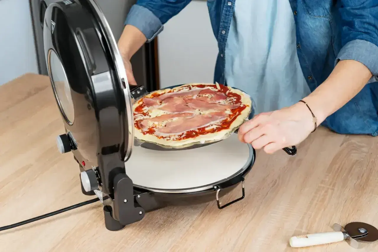 Osoba wkłada pizzę z szynką do pieca. Szybko i łatwo przygotujesz idealną pizzę w domu.