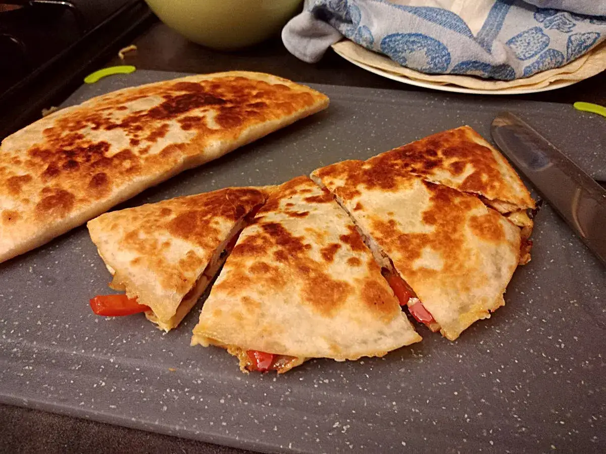 Złocista quesadilla z kurczakiem, pokrojona na trójkąty, leży na desce. Widać kawałki papryki i sera.