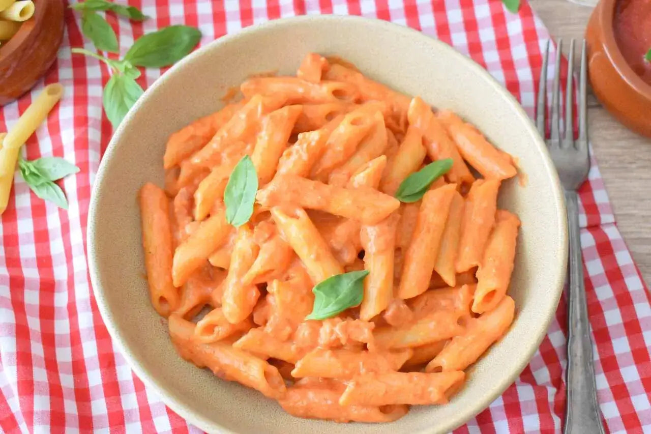 Penne alla vodka w kremowym sosie pomidorowym, udekorowane listkami bazylii, podane na talerzu w kratkę.