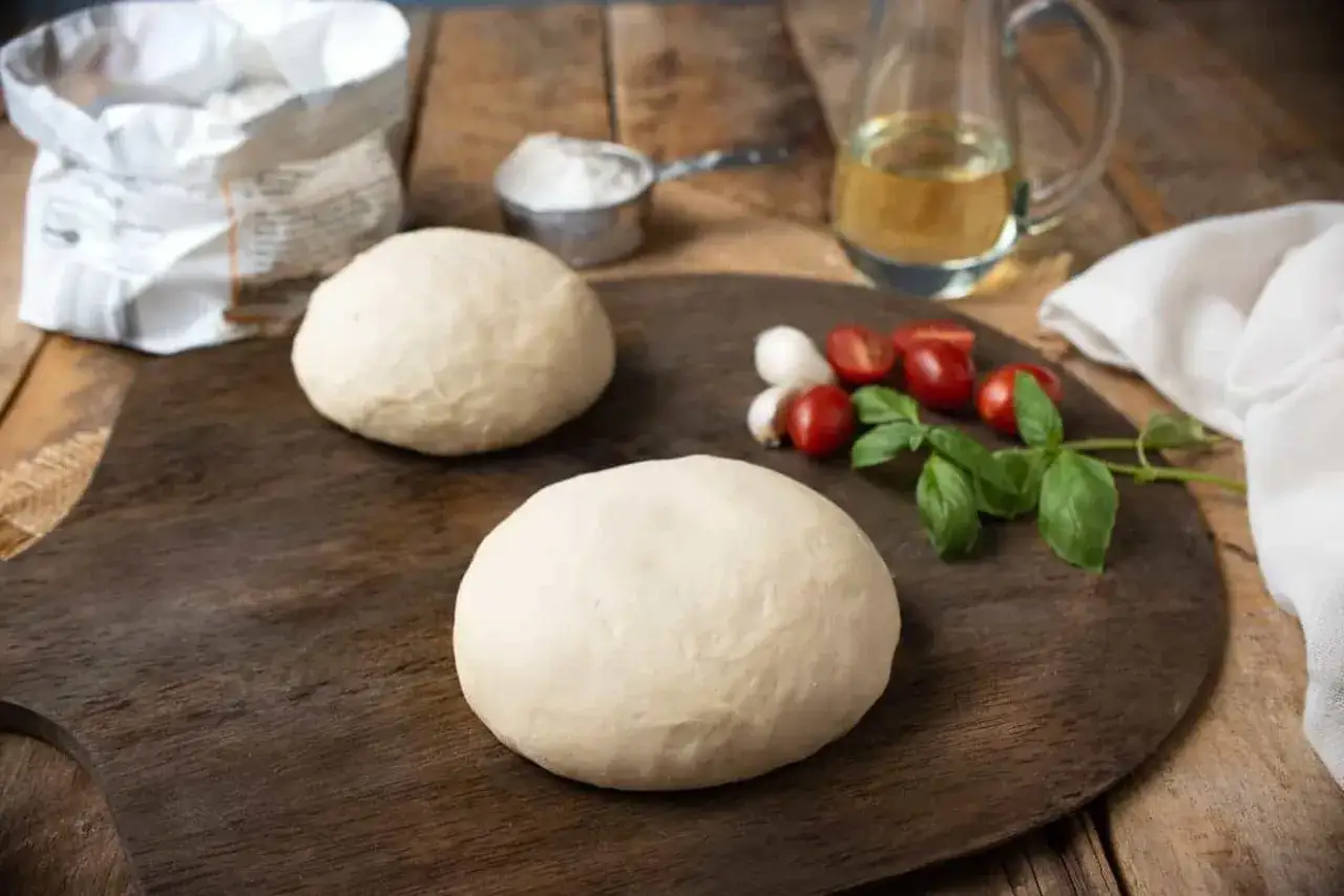 Dwa okrągłe ciasta na pizzę bez drożdży, gotowe do pieczenia. Obok pomidorki, bazylia i oliwa.