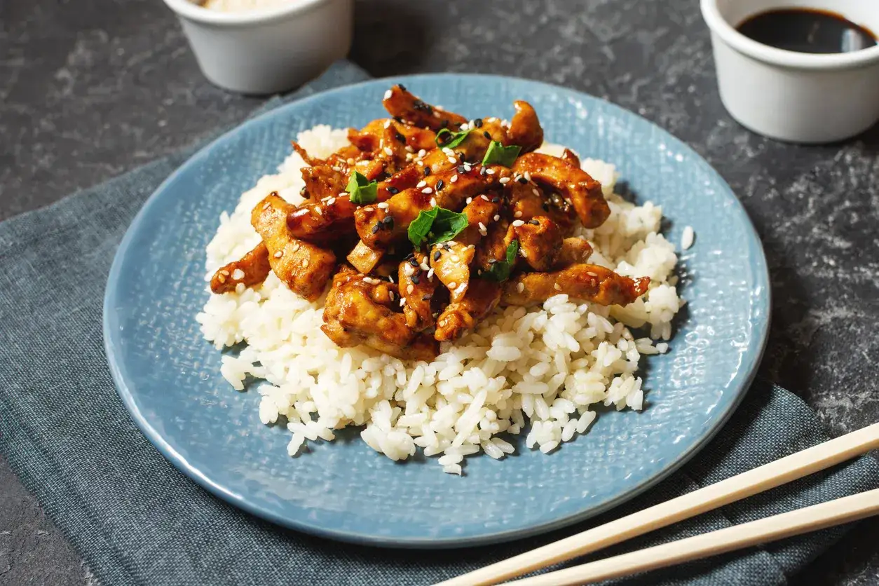 Pyszny kurczak w sosie teriyaki, podany na białym ryżu, posypany sezamem i zieloną pietruszką. Idealny posiłek.