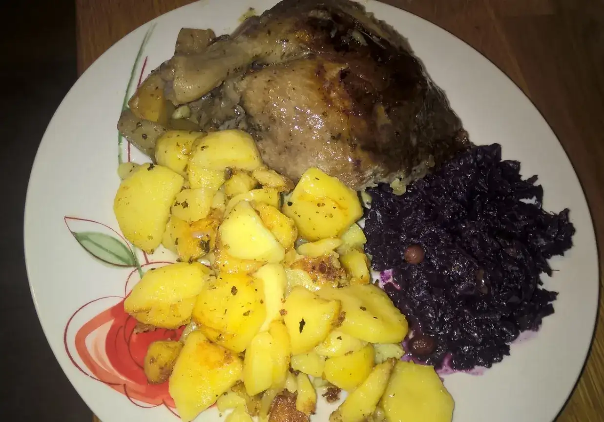 Pieczona kaczka z jabłkami, ziemniakami i czerwoną kapustą na talerzu z kwiatowym wzorem.