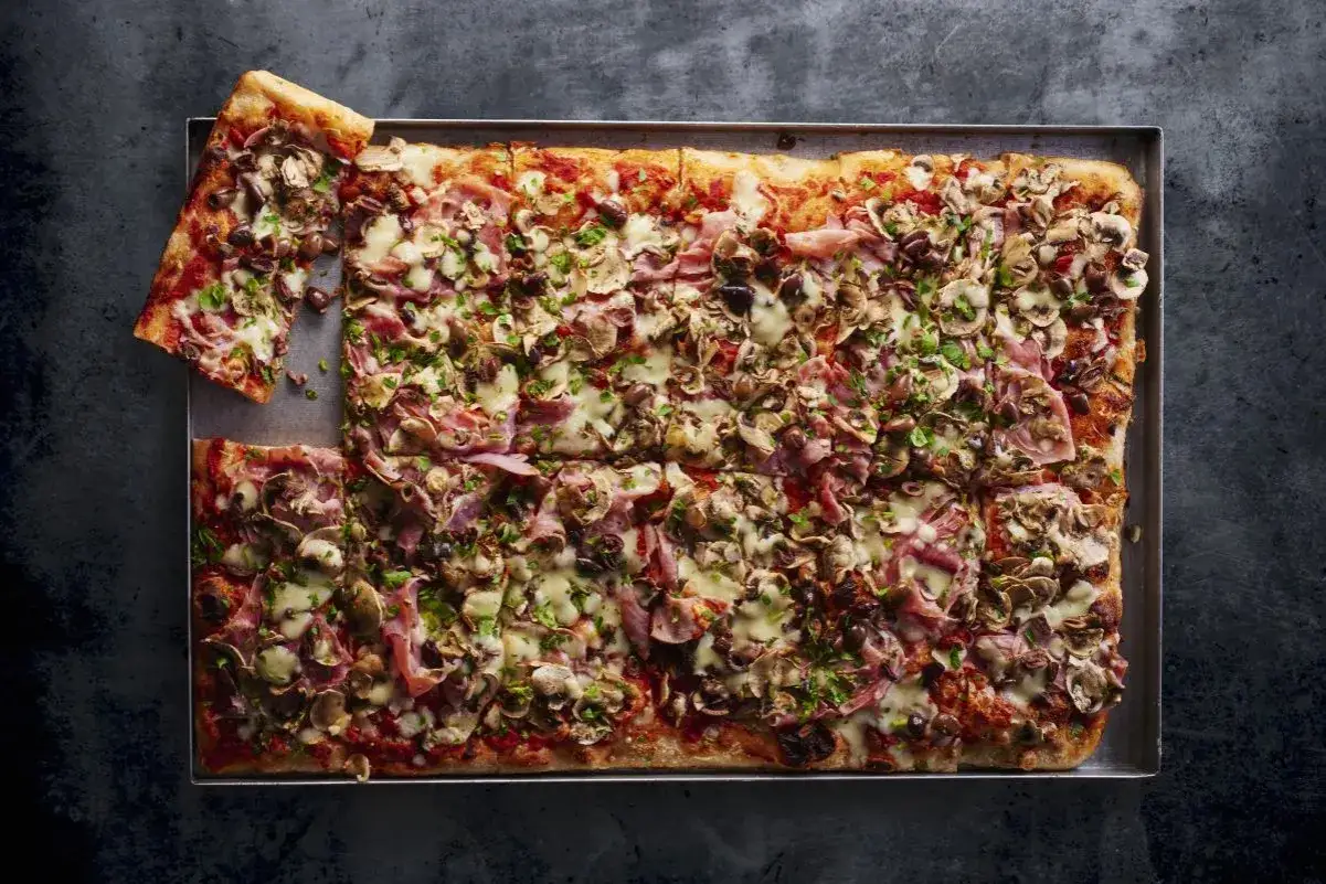 Pyszna pizza domowa przepis na blachę z szynką, pieczarkami i oliwkami, posypana świeżą natką pietruszki.