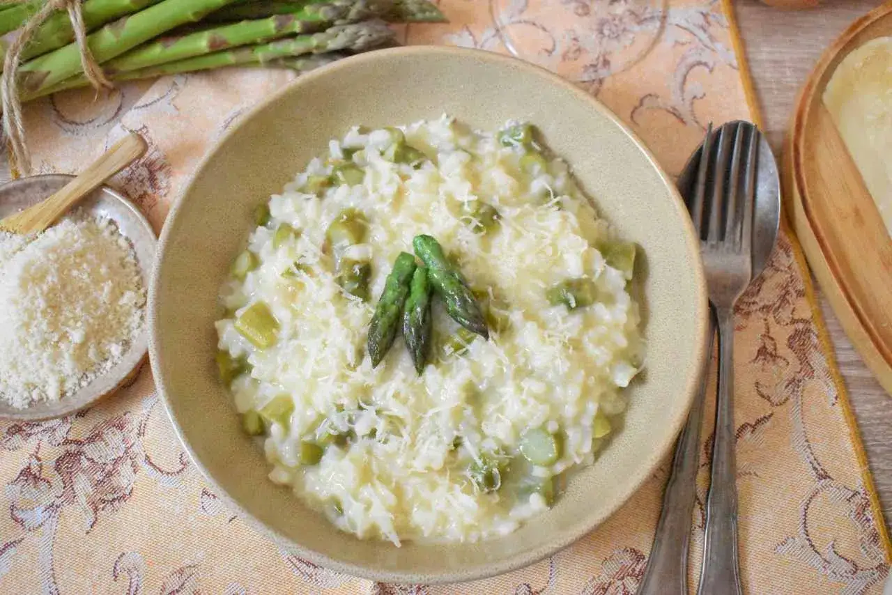 Kremowe risotto ze szparagami, posypane parmezanem, z trzema młodymi szparagami na wierzchu. Obok miseczka z tartym serem i pęczek świeżych szparag&oacute;w.