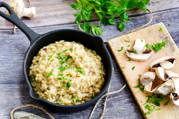 Kremowe risotto z warzywami, posypane natką pietruszki, obok świeże pieczarki na desce.